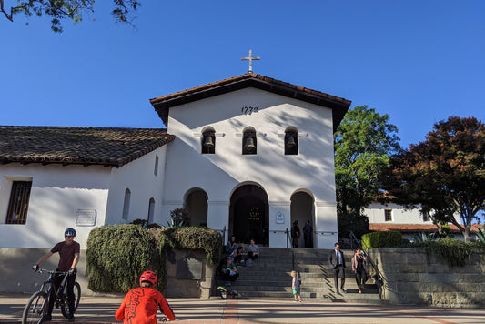 The white bell-tower façade of Mission San Luis Obispo de Tolosa with cyclists passing on a sunny day